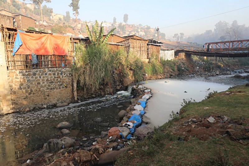 A stretch of the Akaki river with water that appears almost white due to contamination. Several stone and wooden structures, and a bridge, line the riverbank.
